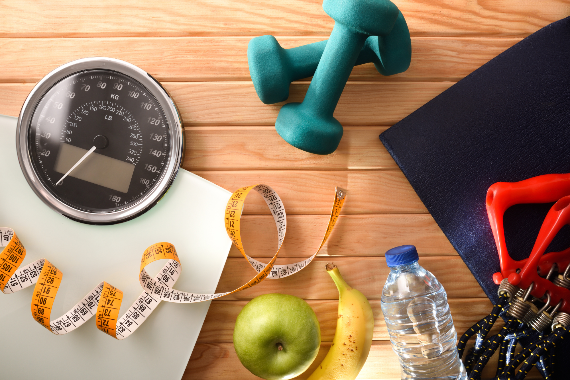 A scale, dumbbells, measuring tape, water bottle, resistance bands, apple, and banana placed on a wooden surface.