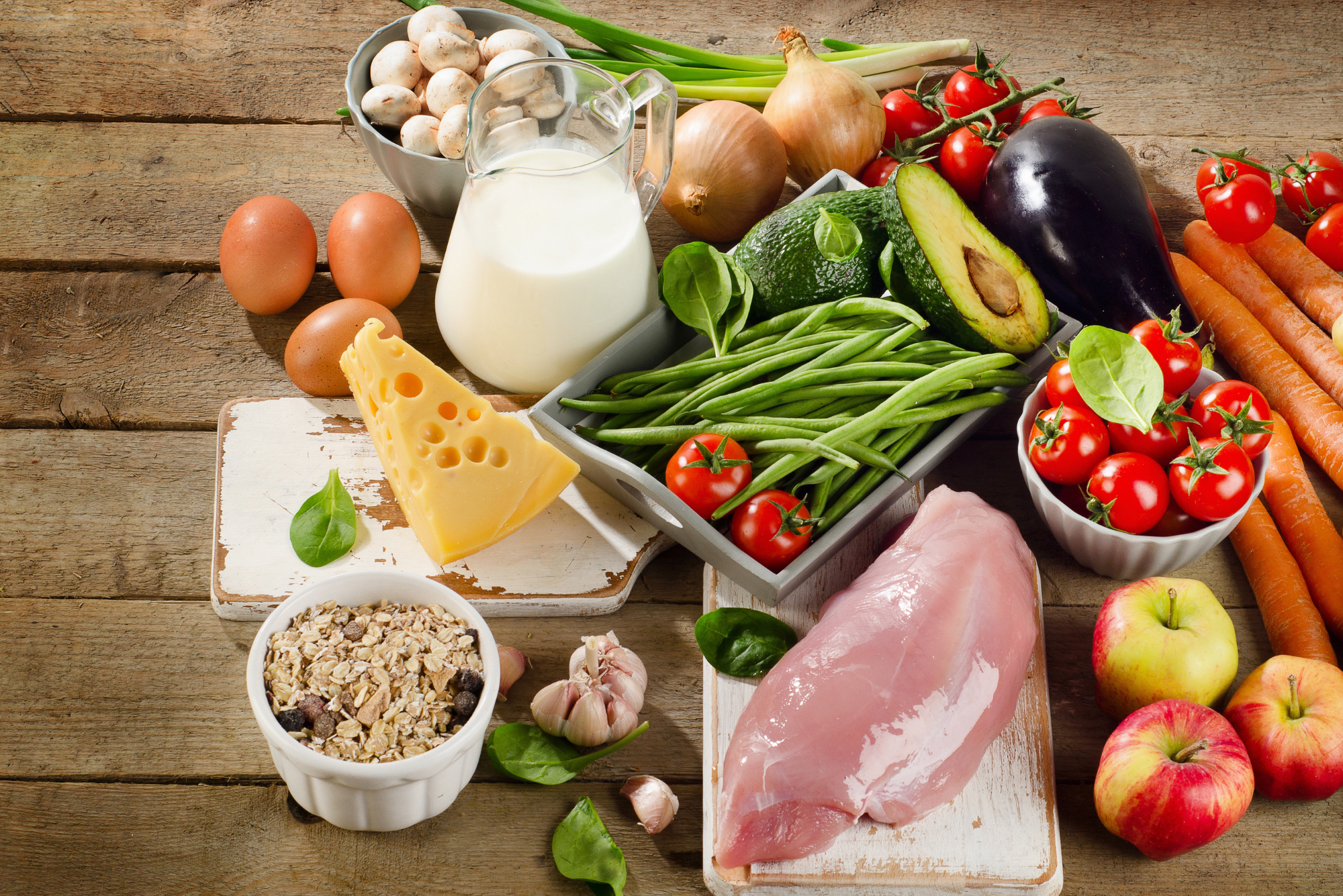 An assortment of fresh foods: chicken, eggs, cheese, milk, vegetables, grains, and fruits on a rustic wooden surface.