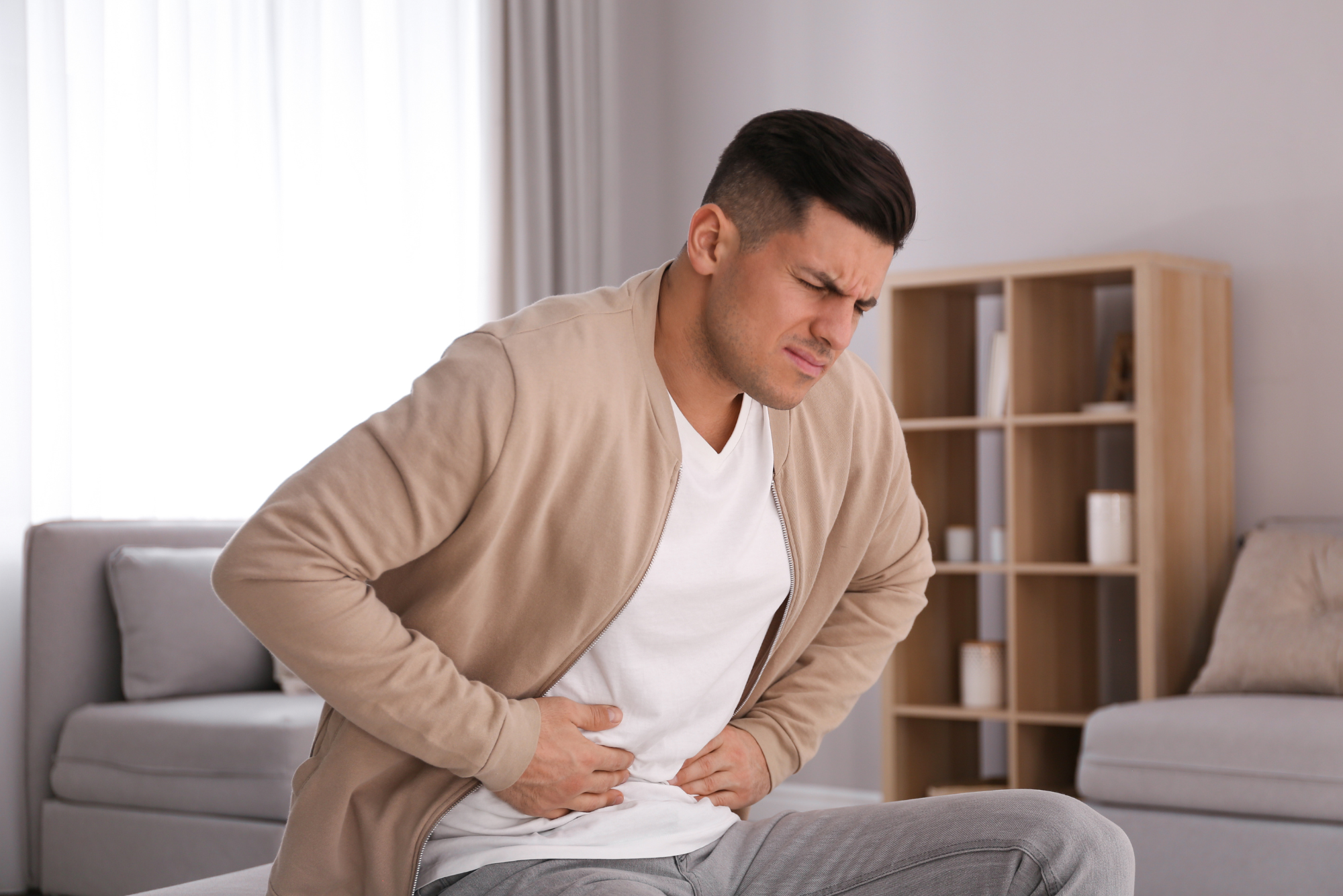 Man grimacing and holding stomach in pain while sitting on couch at home, appearing to have abdominal discomfort.