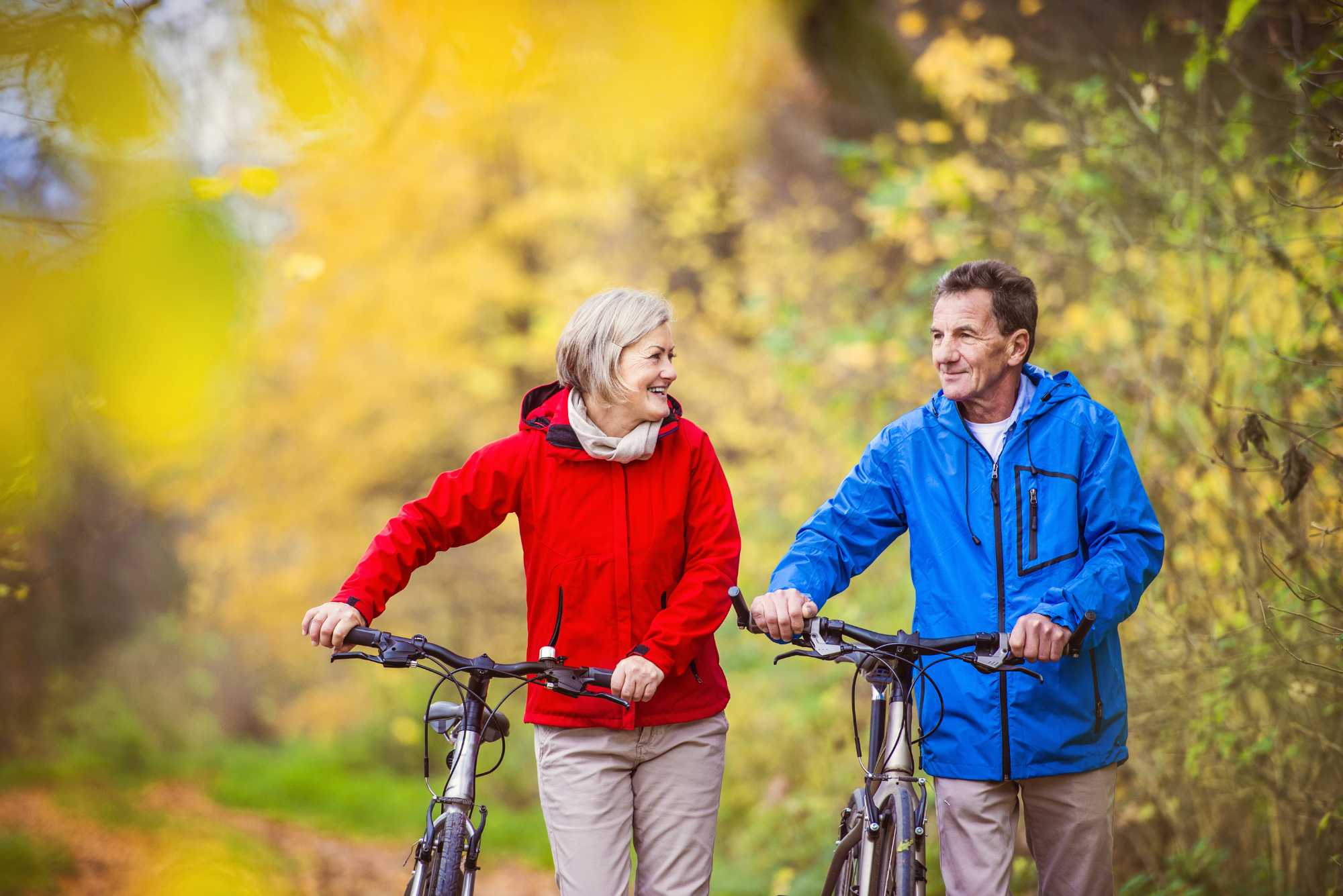 Older couple walking bikes through a sunny, leafy path while smiling and enjoying outdoor activities for stress management.