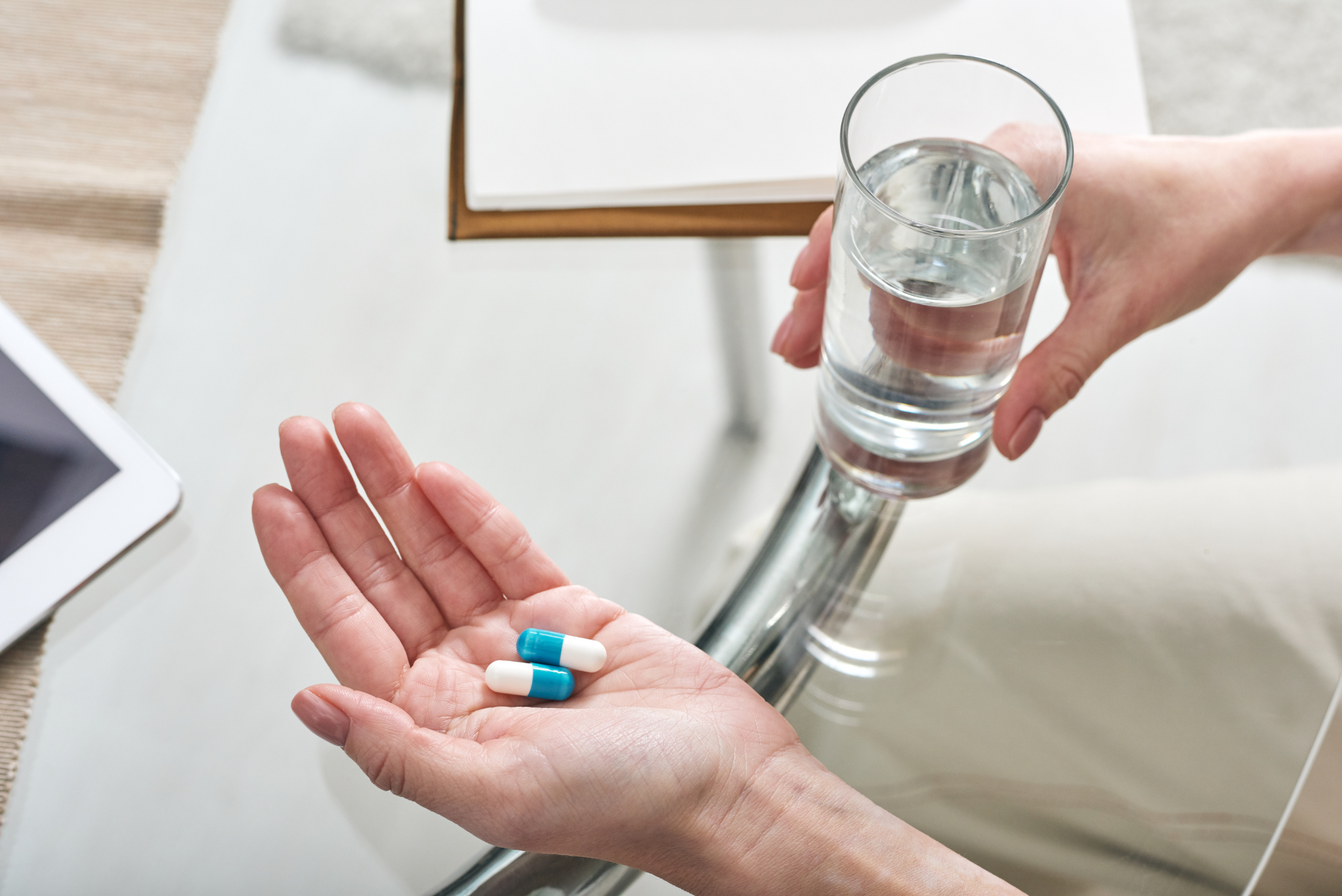 A person’s hand is shown holding two white and blue capsules or pills while the other hand is holding a glass of water.