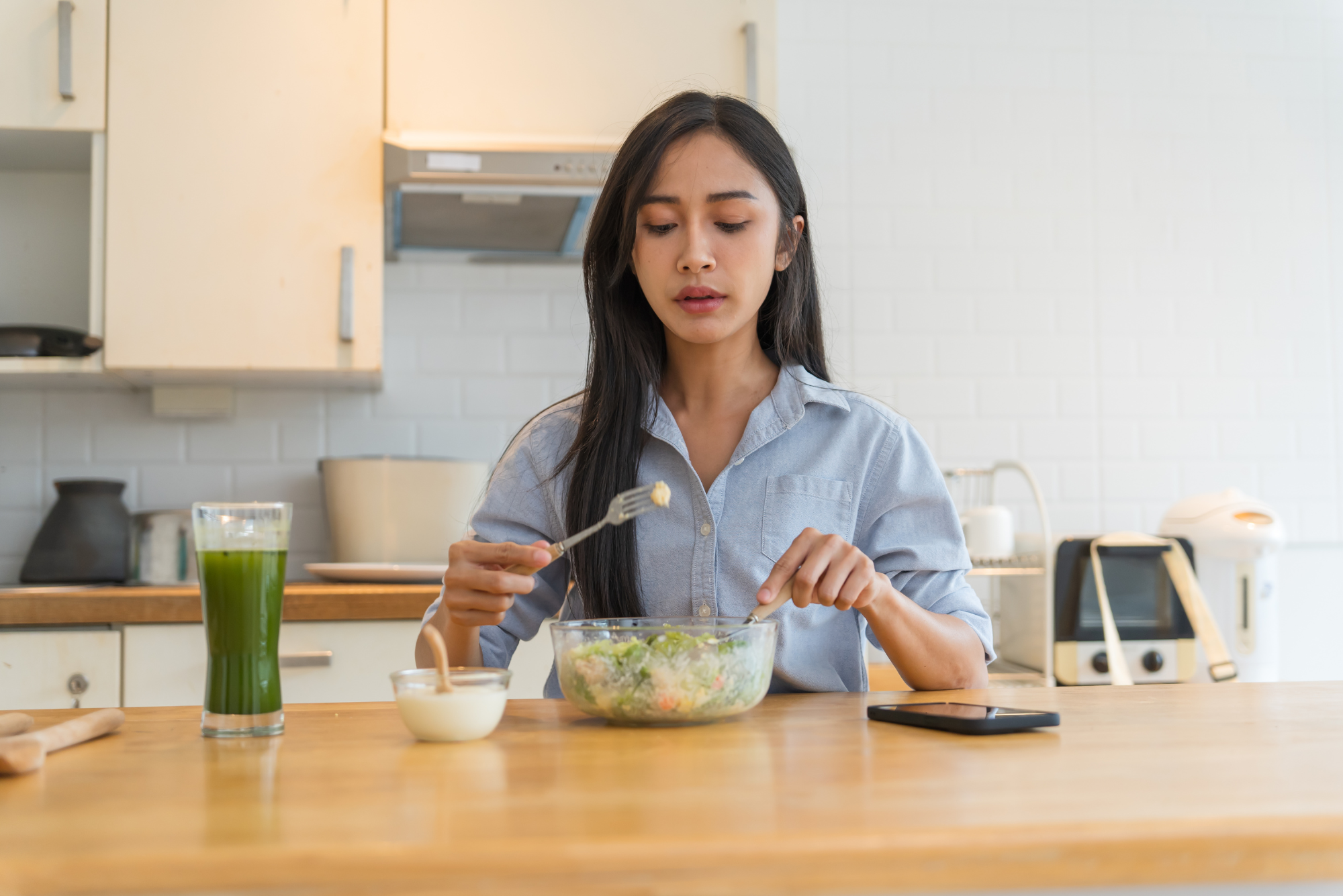 Woman eating healthy salad and green smoothie at kitchen counter, demonstrating mindful eating and meal variation.